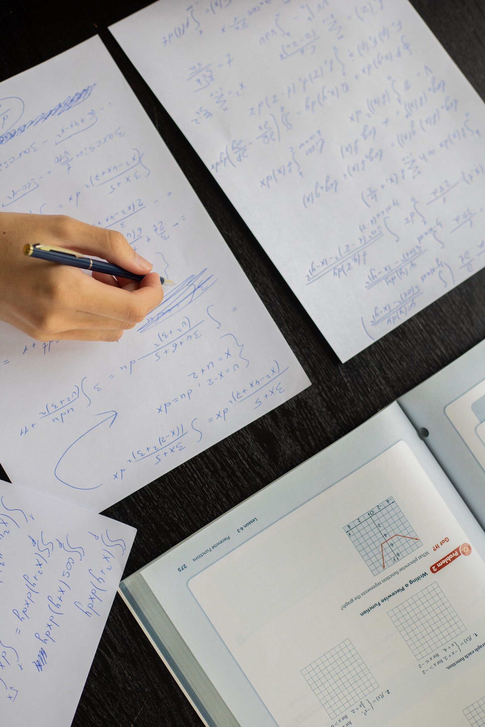 Top view of crop anonymous learner with pen doing mathematics calculations on paper sheet with opened textbook